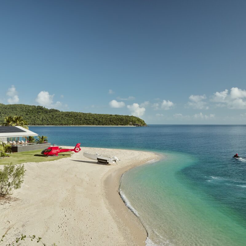 Image showing island with white sand and blue water, helicopter on the beach, jet skis in water, home in background,